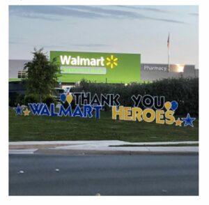 A picture of a Walmart in the background, painted green with white text. In the foreground there is a sign put on a lawn that says "Thank You Walmart Heroes"