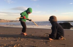 two kids playing on the beach. One is wearing a green sweater and carrying a stick. The other is crouched and wearing a blue sweatshirt
