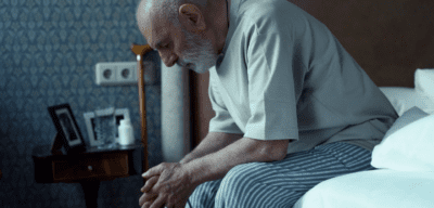 An elderly man sits on the edge of his bed looking forlorn. In the background is a cane and some medicine bottles.