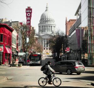 A Black man rides a bicycle in the business district in Madison, Wisconsin. In the background is the state capitol building.