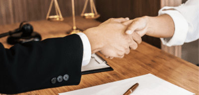 Two people shaking hands. One person is wearing a suit. There is a Scales of Justice in the background.