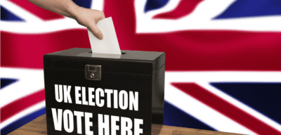 Image of a voting box that says "UK Election: Vote Here." The UK flag is in the background.